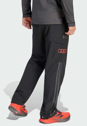 Person wearing black pants with side pocket and red Audi rings logo, paired with bright red and black athletic shoes.