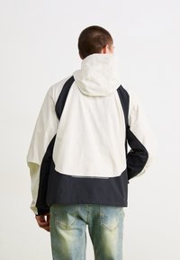 Hettejakke med en crème-farget front og svart bak, laget av et lett materiale med en strømlinjeformet design og en løs passform.