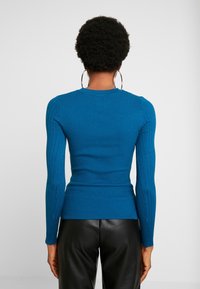 Haut à manches longues côtelé en bleu vif, avec une silhouette ajustée et un col rond. La texture est lisse avec un détail côtelé sur l'ensemble.