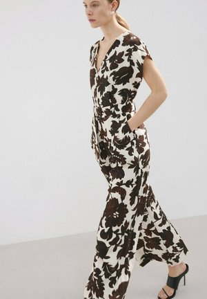 Mujer vestida con un mono blanco con un gran estampado floral marrón oscuro, bolsillos laterales y sandalias negras de tacón alto, caminando frente a un fondo liso.