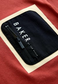 Roter Stoff mit einer schwarzen Reißverschlusstasche und einem cremefarbenen rechteckigen Rand; der Text lautet "BAKER by TED BAKER" in Weiß.