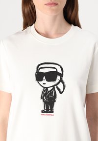Balta medvilninė marškinėlių su juodu komiksiniu piešiniu, kuriame pavaizduotas vyras akinių nuo saulės ir kostiumo, o apačioje užrašyta "KARL LAGERFELD".