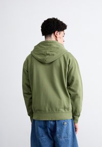 Olive green hoodie with a front pocket, featuring a drawstring hood and ribbed cuffs. Matched with blue denim pants. Smooth texture.