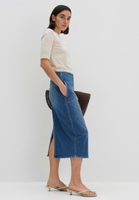 Jupe en denim bleu moyen avec des fentes latérales, associée à un haut crème ajusté. La modèle tient un sac à main marron et porte des sandales beige à bout ouvert.