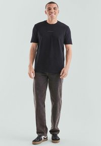 Schwarzes Baumwoll-T-Shirt mit Rundhalsausschnitt und kurzen Ärmeln, versehen mit einem kleinen roten Textlogo, kombiniert mit dunkelbraunen Jeans und schwarzen Turnschuhen.