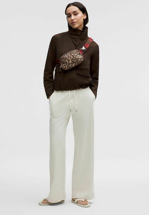 Femme portant une veste marron foncé, un pantalon blanc à jambes larges, des tongs et un sac bandoulière imprimé léopard avec une sangle rouge, debout les mains dans les poches.