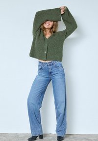 Olivu zaļš īss kardigans ar teksturētu adījumu, pogu aizdare, kombinēts ar augsto jostu ziliem džinsiem un melniem potīšu zābakiem.