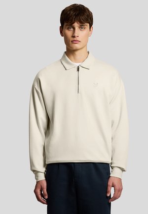 Junger Mann mit braunen Haaren, der einen beigen Pullover mit langen Ärmeln und Reißverschluss im Viertelmaß sowie eine marineblaue Hose trägt und vor einem schlichten grauen Hintergrund steht.