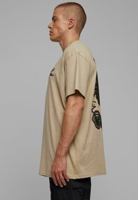 Beige cotton t-shirt with short sleeves, featuring a graphic print of a pharaoh on the back, with a loose fit and slightly elongated shape.