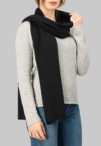 Sorte uldhalstørklæde draperet omkring halsen, der kontrasterer med en lysegrå sweater. Halstørklædet har en blød tekstur og en rektangulær form.