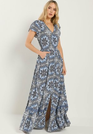 Vrouw die een maxi-jurk draagt met een blauw-wit bloemenpatroon, korte mouwen en een split aan de voorkant, staand met één hand in een zak.