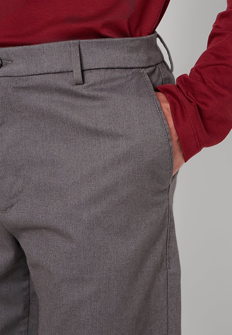 Graue, strukturierte Hosen mit schmaler Passform, Taillenbund mit Gürtelschlaufen und seitlichen Taschen. Kombiniert mit einem langen rotärmeligen Hemd.