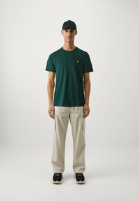 T-shirt verde in cotone con un piccolo logo giallo, abbinata a pantaloni cargo beige chiaro con tasche laterali e scarpe sportive nere.