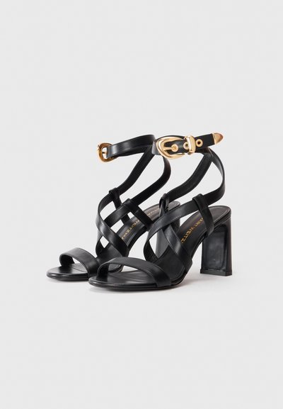 Zwarte leren hoge hak sandalen met kruislings straps en gouden gespsandalen rond de enkels tegen een effen witte achtergrond.