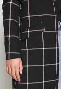 Main reposée sur une veste noire à motif en quadrillage blanc et poche à rabat, à côté d'un jean en denim bleu.