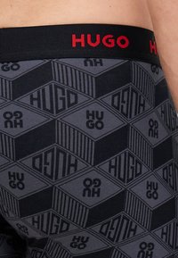 Boxers cortos grises para hombre con un patrón geométrico repetido. Cuenta con una cinturilla elástica negra con la palabra "HUGO" en letras rojas. Textura de tela suave.