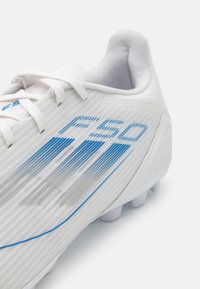 Scarpone da calcio bianco con superficie texturizzata, accenti e grafiche blu, e lacci bianchi. Presenta una forma curvata e distintivi tacchetti sulla suola.
