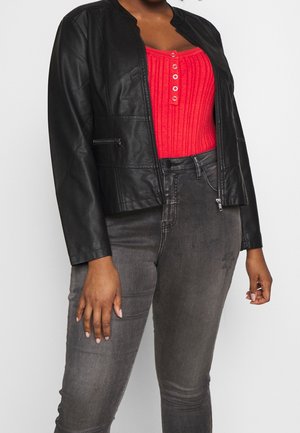 Personne portant une veste noire en simili cuir, un haut rouge côtelé à boutons et un jean gris taille haute, debout devant un fond uni.