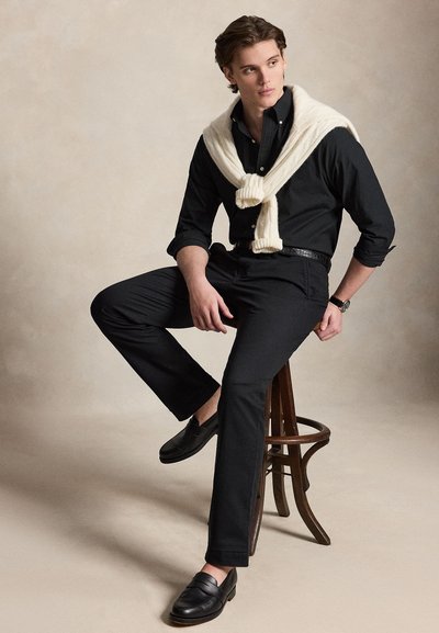 Νεαρός άνδρας που κάθεται σε ένα ξύλινο σκαμπό, ντυμένος ολόκληρος με μαύρα ρούχα, με ένα λευκό πουλόβερ περασμένο στους ώμους, κοιτάζει προς τα πλάγια.