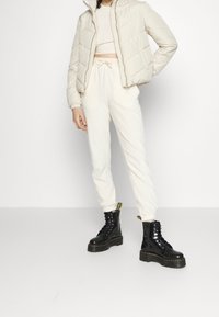 Beżowa pikowana kurtka puchowa, krótki beżowy top, kremowe dresowe spodnie i czarne sznurowane masywne botki z żółtymi akcentami. Minimalistyczny look.