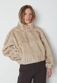 Giacca in pelliccia sintetica beige con colletto alto, chiusura con zip, polsini arricciati e orlo, con un accento metallico all'collo.