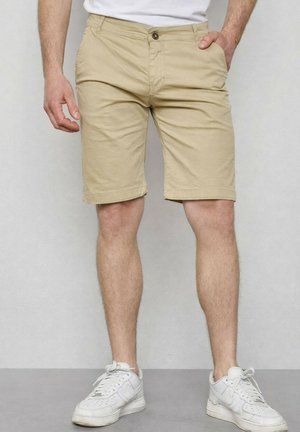 Homme portant un short beige jusqu'aux genoux et des baskets blanches, se tenant devant un fond gris uni.