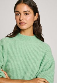 Mintgroene geribbelde trui met een hoge hals, gemaakt van zachte stof. Model poseert met gekruiste armen, waarbij de textuur en kleur worden getoond.