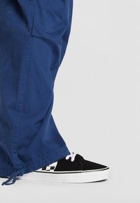 Svarte semskede sneakers med hvite snørebånd og rutete detaljer, kombinert med løstsittende blå bukser med snøring i hemmen.
