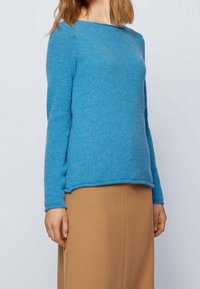 Femme portant un pull en tricot bleu à manches longues et une jupe camel taille haute sur un fonds clair.