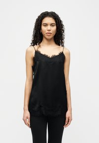 Νεαρή γυναίκα με σγουρά μαλλιά, φορώντας μαύρο καμιζολάκι με δαντέλα και μαύρο παντελόνι, στέκεται μπροστά σε απλό λευκό φόντο.