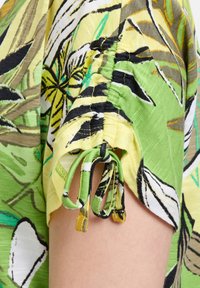Bluzka z krótkim rękawem w zielono-żółtej tkaninie z kwiatowym wzorem, z marszczonymi rękawami z wiązanymi akcentami oraz lekką teksturą.