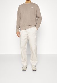 Beige sweatshirt med rund halsringning och vit logotyp, kombinerad med ljusbeiga byxor och beige sneakers med ett distinkt mönster.