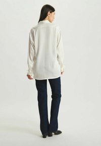 Chemise blanche à manches longues avec une coupe décontractée, poignets boutonnés et empiècement au dos. Associée à un jean évasé bleu foncé et des chaussures noires.