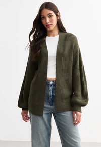 Olive green knitted cardigan with wide sleeves, open front, textured ribbing, worn over a white top and high-waisted denim jeans.