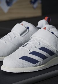 White athletic shoes with a textured finish, featuring three dark blue stripes, a white Velcro strap, and orange detailing on the heels.