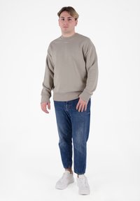 Maglione beige con collo rotondo, polsini e orlo a coste, abbinato a jeans blu tagliati e sneakers bianche. Dettaglio di logo semplice sul petto.