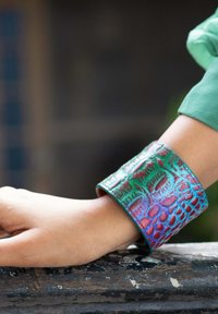 Armreif aus strukturiertem Leder in schillerndem Grün und Rot, mit einem reptilienartigen Muster und glatten Kanten, am Handgelenk getragen.