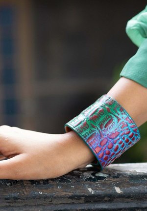 Knooparmband gemaakt van gestructureerd leer in iriserend groen en rood, met een reptielenpatroon en gladde randen, gedragen om een pols.