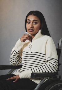 Maglione bianco a costine con strisce orizzontali nere, colletto alto e dettaglio con zip. Modello seduto su una sedia a rotelle, indossando pantaloni scuri.
