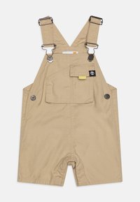 Salopette en coton beige clair avec des bretelles réglables, des accents de boutons-pression, une poche avant et une coupe décontractée, dotée d'un design de jambes ouvertes.