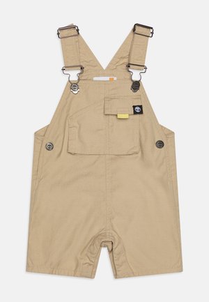 Salopette en coton beige clair avec des bretelles réglables, des accents de boutons-pression, une poche avant et une coupe décontractée, dotée d'un design de jambes ouvertes.