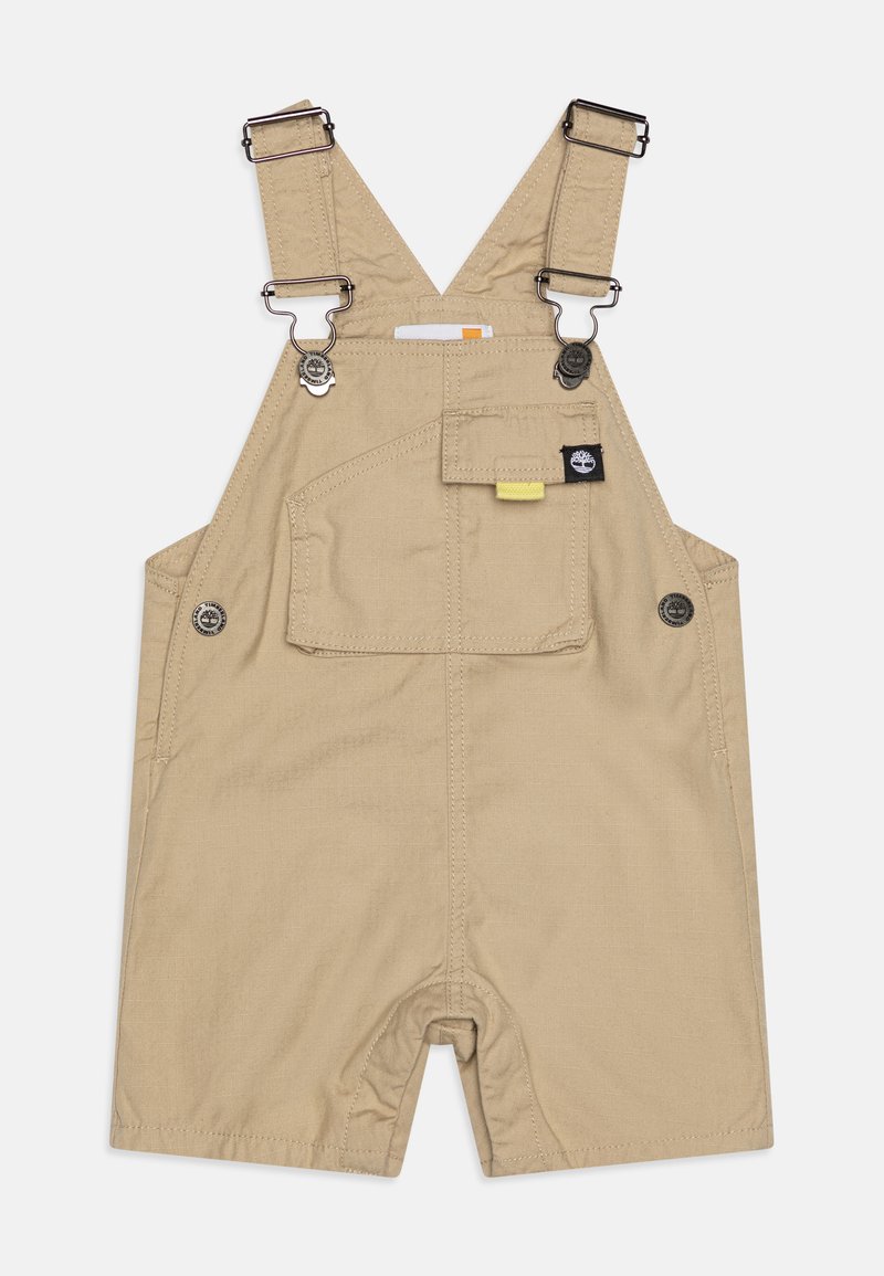 Salopette en coton beige clair avec des bretelles réglables, des accents de boutons-pression, une poche avant et une coupe décontractée, dotée d'un design de jambes ouvertes.