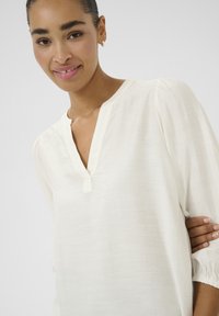 Blouse blanche légère avec un décolleté en V, des manches bouffantes et un motif texturé subtil. Tissu doux et respirant avec une coupe décontractée.