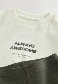 Tricou alb din bumbac cu text negru "ÎNTOTDEAUNA FANTASTIC" și "LOS ANGELES, CALIFORNIA" dedesubt, suprapus peste un material cu dungi verde închis.