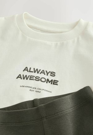 Bílý bavlněný tričko s černým nápisem "VŽDY ÚŽASNÝ" a nápisem "LOS ANGELES, KALIFORNIE" pod ním, položené na tmavě zelené žebrované látce.