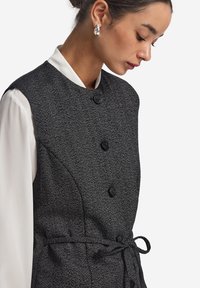 Grå väst med houndstooth-mönster, strukturerad design, svarta knappar och ett bälte i midjan, över en vit blus.