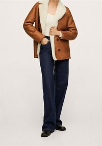 Persona che indossa un giubbotto di shearling marrone, una maglietta bianca, jeans a gamba larga blu scuro e scarpe nere, in piedi con una mano in tasca.