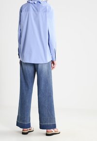 Camicia azzurra a maniche lunghe con colletto arricciato, abbinata a jeans scuri a gamba larga con orli sfilacciati e cuciture bianche.