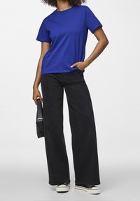 Donna che indossa una maglietta blu, jeans larghi neri e sneakers con platform neri, con una borsa nera al suo fianco.