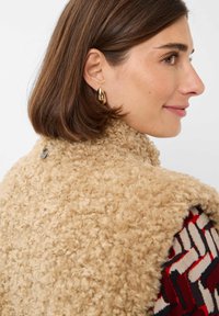 Beige nepbont gilet met een gestructureerde, fluffy afwerking. Heeft een zilveren logo-tag aan de achterkant. Kleurrijke patterndop zichtbaar eronder.